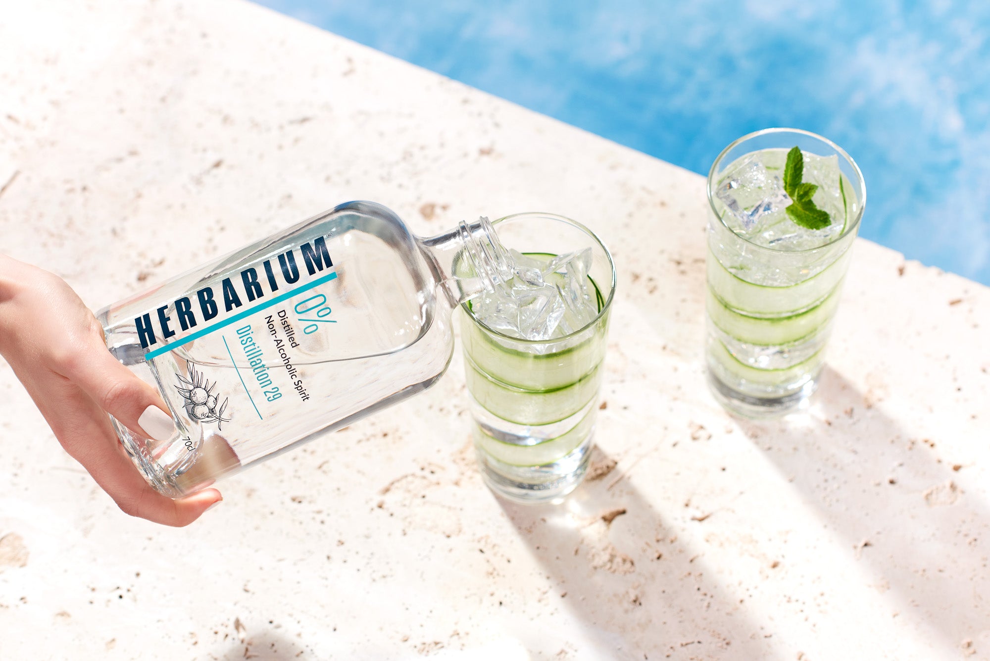 Herbarium Distillation 29 poured into a glass, with ice, cucumber with mint garnish beside swimming pool